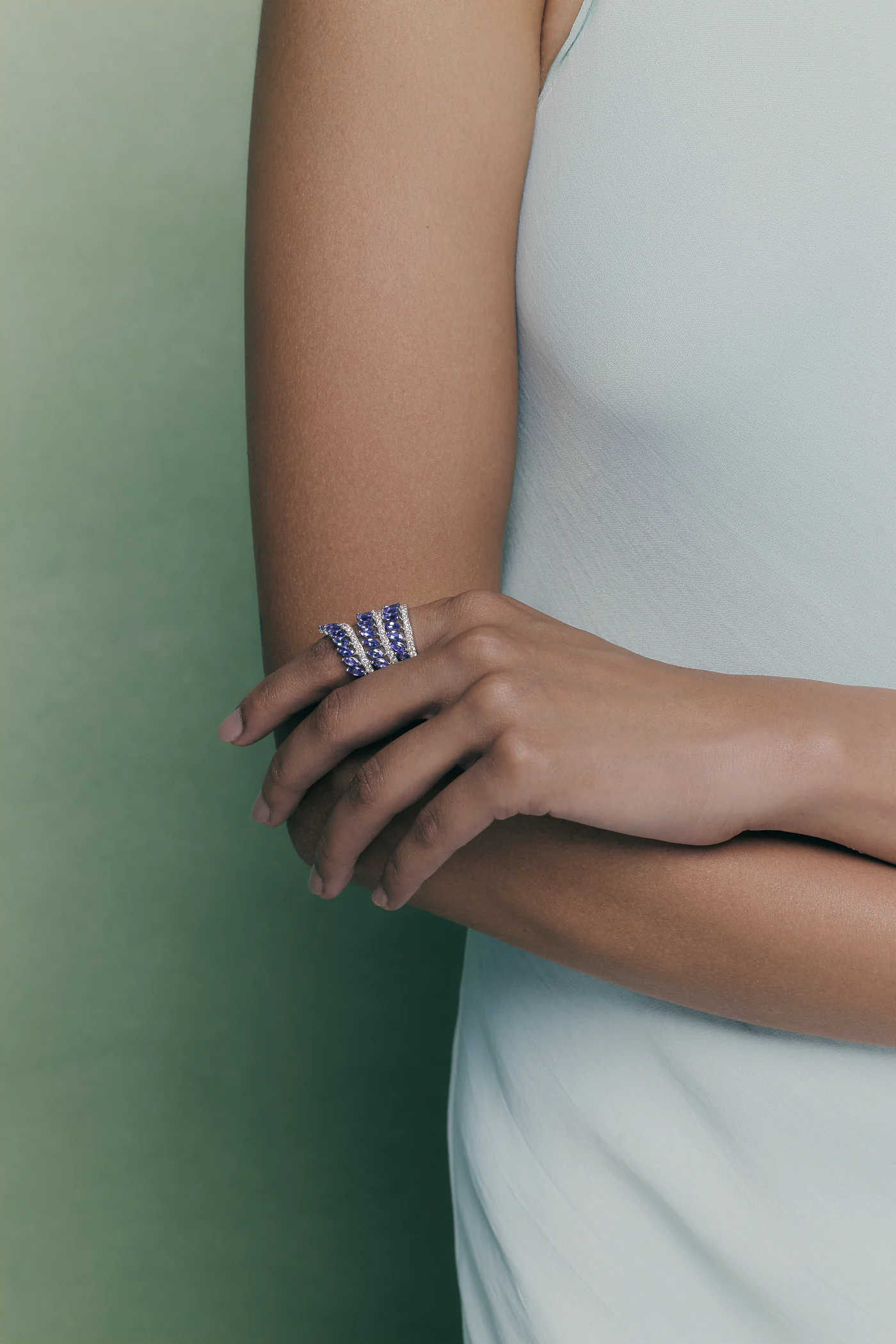 A hand resting on a folded arm, wearing a wide tanzanite and diamond ring against a pale green wall.