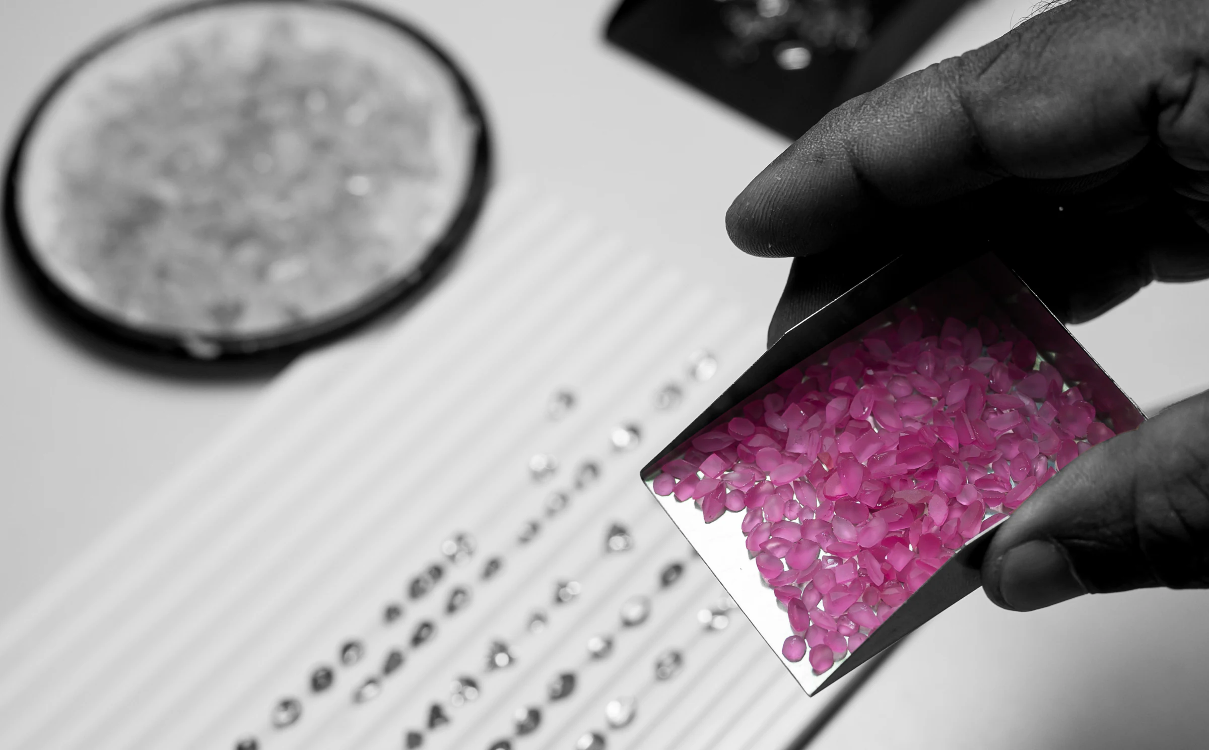 A hand tipping a paper of rough pink spinel beside a grading tray of cut stones.